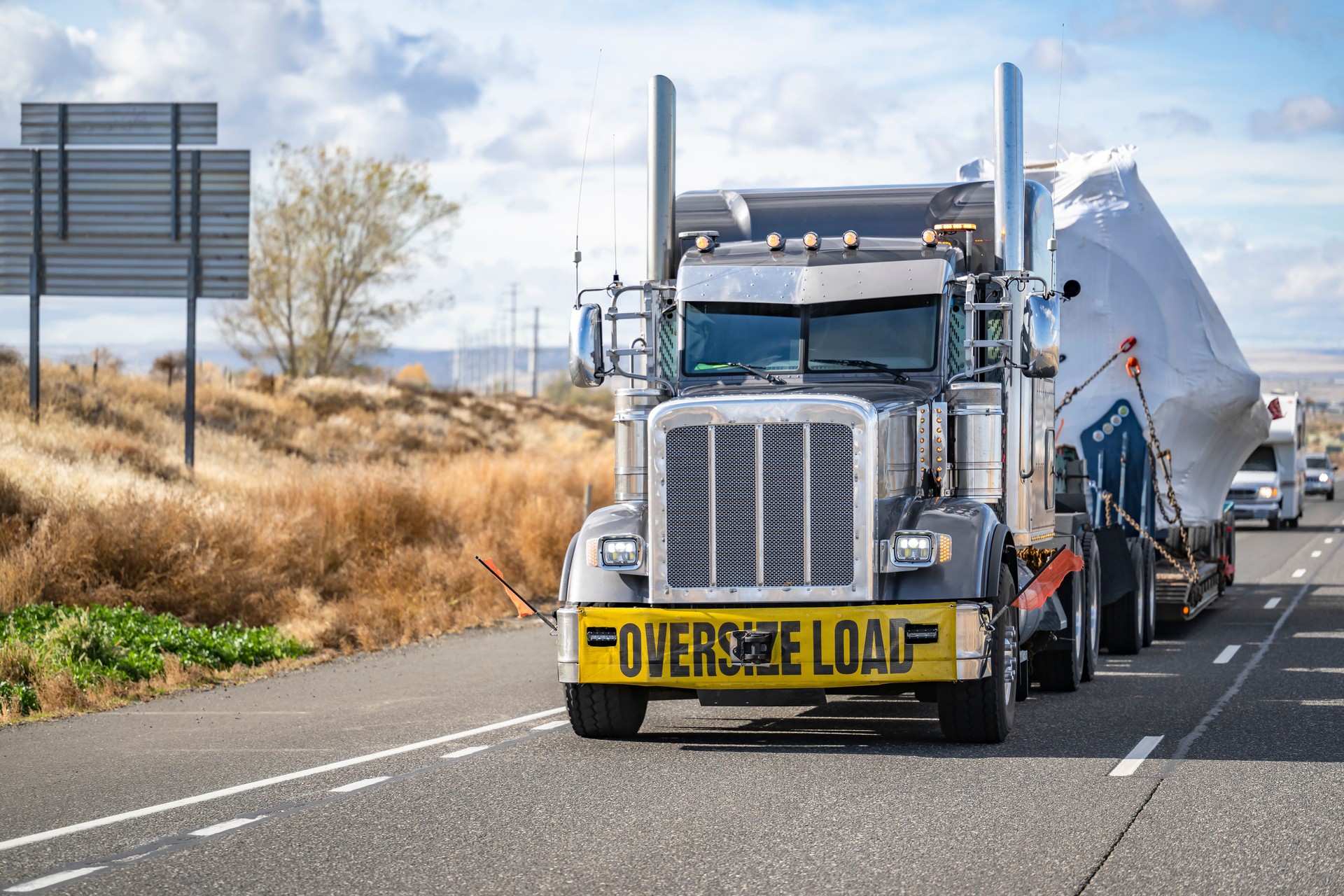 Classic American big rig black semi truck with oversize load sign transporting heavy duty covered oversized cargo on step down semi trailer driving on the highway road in front of another traffic